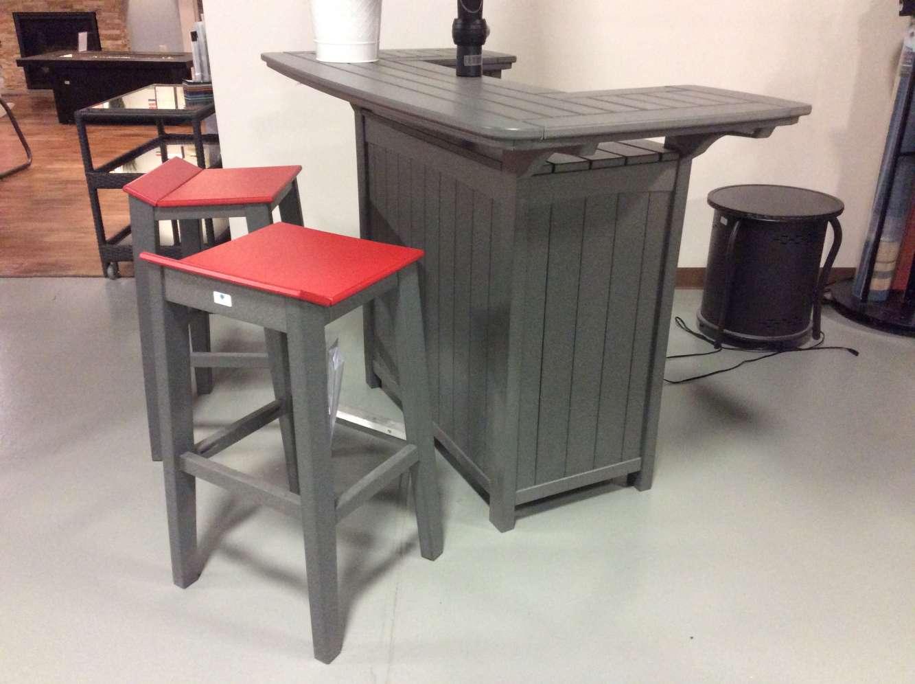 A gray bar-height table with two matching stools that have red cushions, set on a light gray floor beside a sleek insert fireplace in an indoor setting.