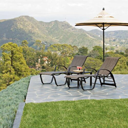 Two lounge chairs and a fire pit under an umbrella on a stone patio, overlooking lush green mountains and trees under a clear sky.