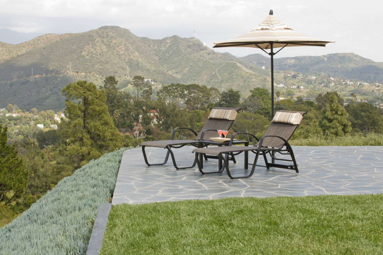 Two lounge chairs and a fire pit under a patio umbrella on a tiled deck with a panoramic view of lush, green hills under a cloudy sky.