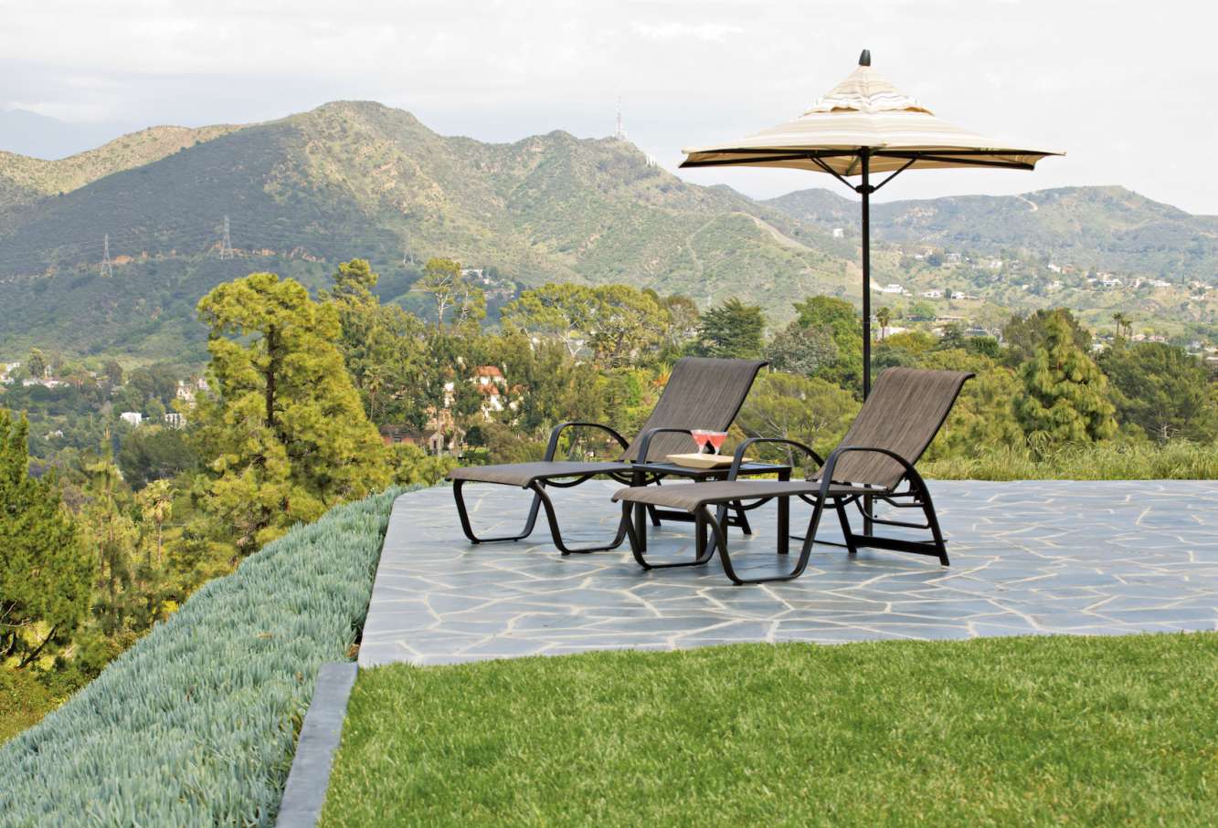 Two lounge chairs and a fire pit under an umbrella on a stone patio, overlooking lush green mountains and trees under a clear sky.