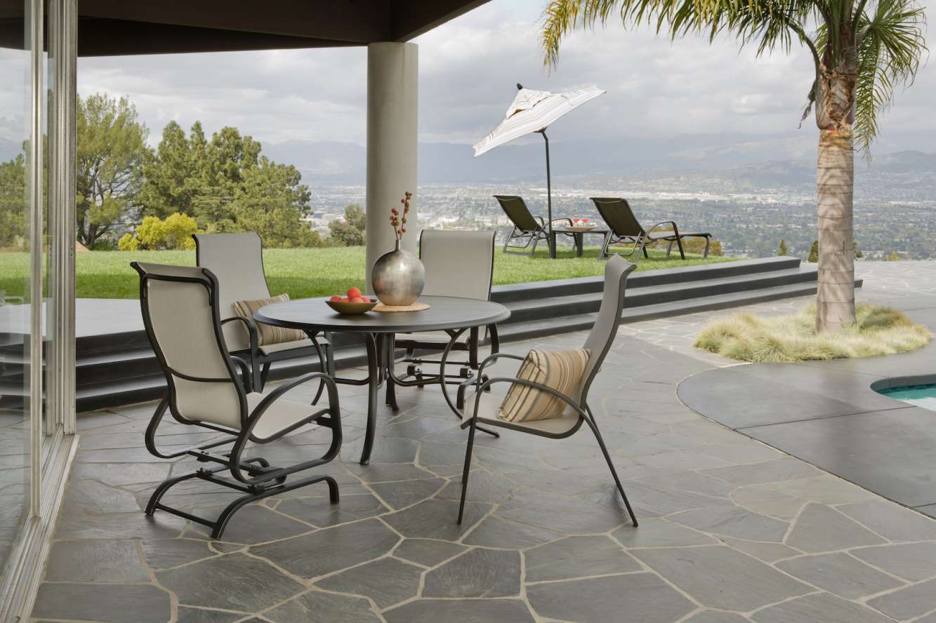 Outdoor patio with a table set and four chairs under an open white umbrella, overlooking a scenic view of mountains and a lush valley. Nearby, two lounge chairs beside a pool feature an insert fire pit for