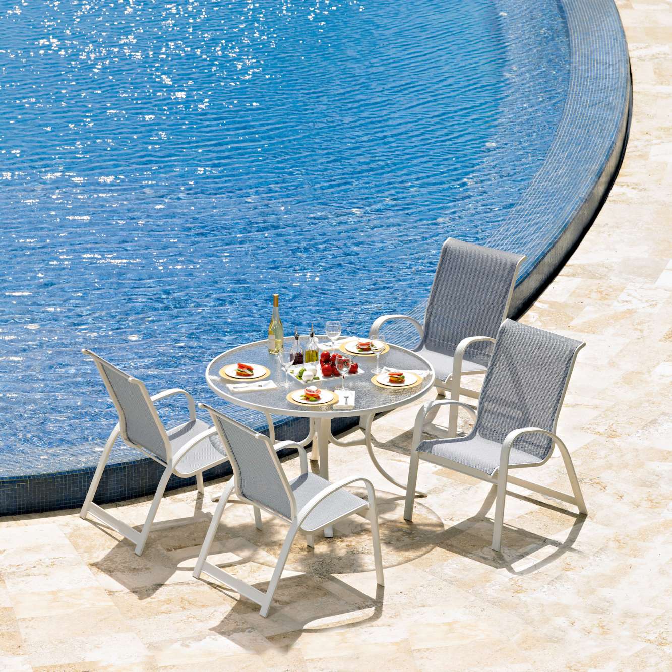 A lunch setup for four people next to a curvy outdoor pool and a fire pit on a sunny day, including plates of food, glasses, and a bottle of champagne.