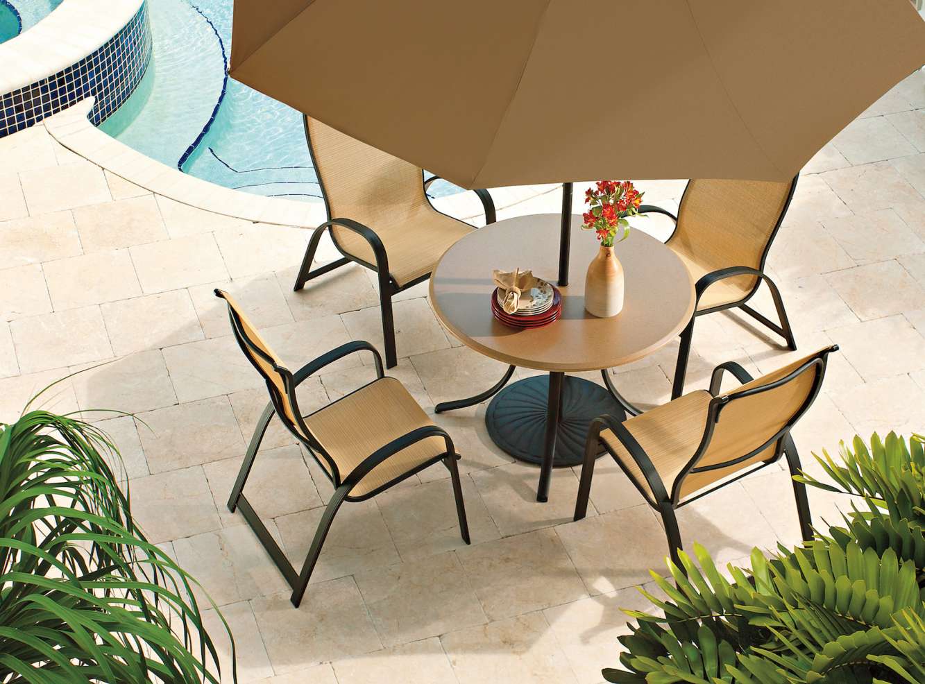 Outdoor patio setting with a table and four chairs under a large umbrella beside a swimming pool, surrounded by lush greenery. A book and a vase with red flowers are on the table, adjacent to an