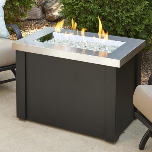 A modern outdoor gas fire pit with a square design, featuring a dark metallic frame and clear fire glass, with yellow flames, set in a patio environment with garden chairs nearby.