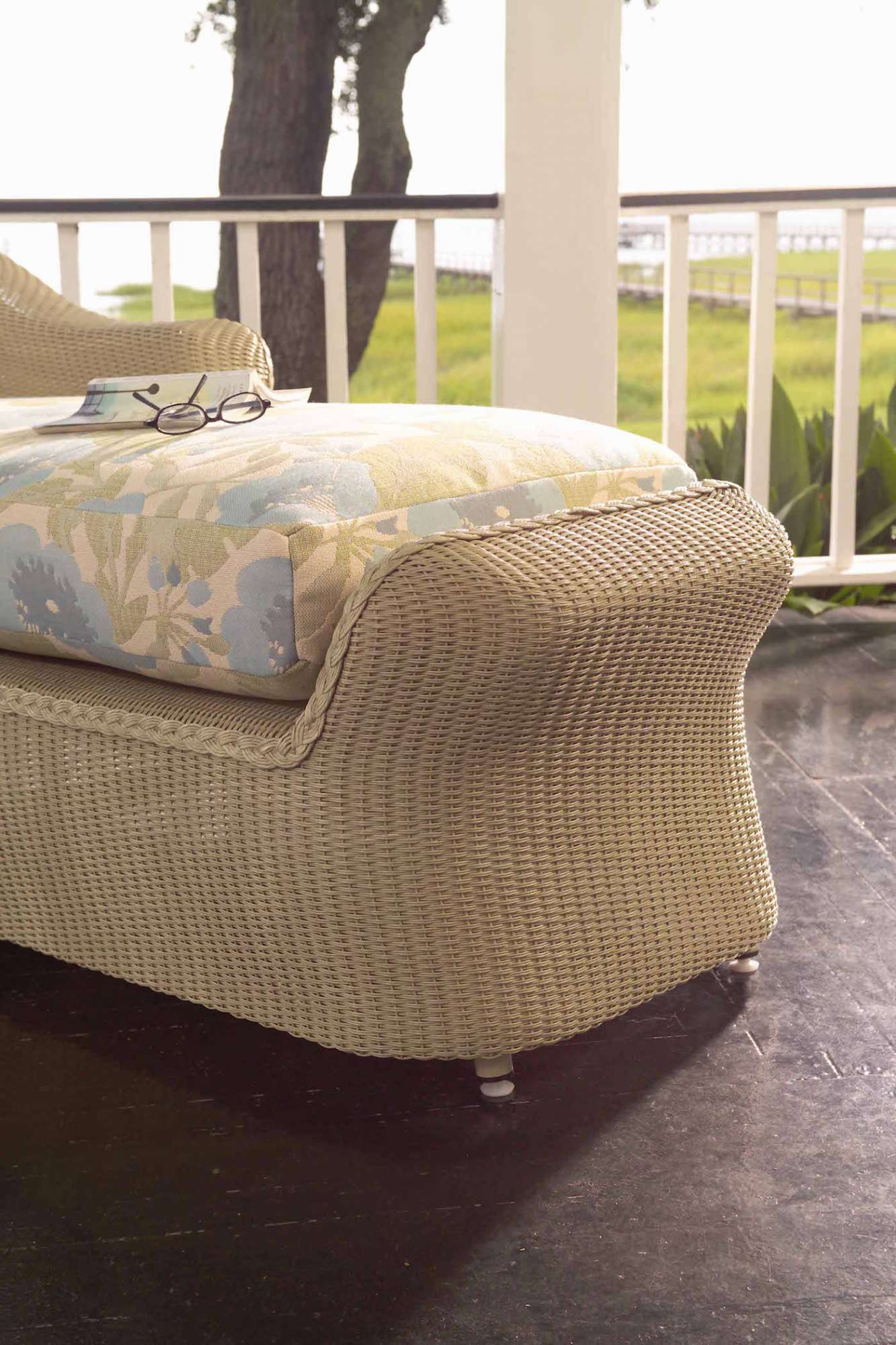 A cozy wicker sofa with floral cushions on a porch overlooking a grassy yard enclosed by a white railing, suggesting a relaxing outdoor space with an inserted fire pit.