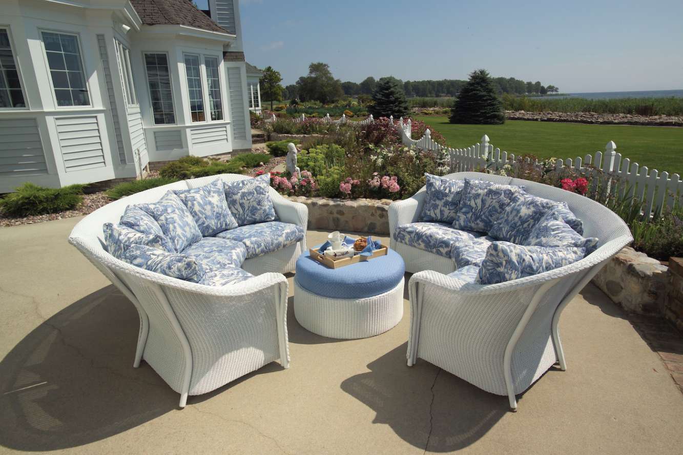 Outdoor patio furniture with wicker sofas and blue cushions set on a stone pavement with a lush garden and lake view in the background. A table holds a tray with refreshments, and nearby, a fire