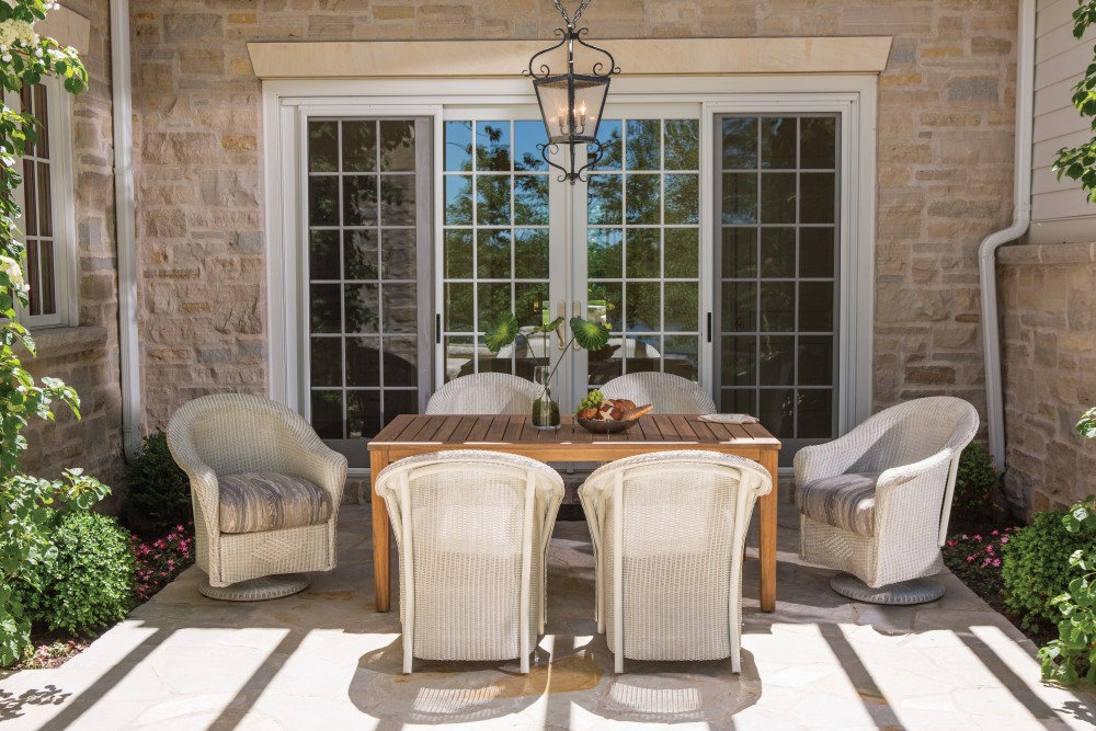 A cozy outdoor dining area with a wooden table and four wicker chairs on a patio, framed by a stone house and glass doors under a hanging lantern. A fire pit insert enhances the scene, bright