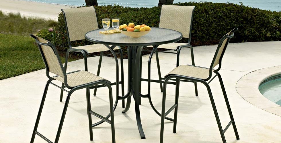 An outdoor patio set with four chairs and a round table, featuring refreshments and fruit, located beside a pool with a fire pit and overlooking a grassy area with a beach view.
