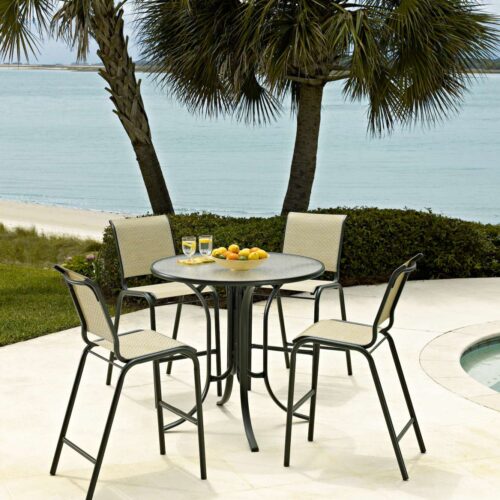 An outdoor dining setup near a pool, featuring a metal table with four chairs and fruit on the table, with palm trees and a fire pit in the background.