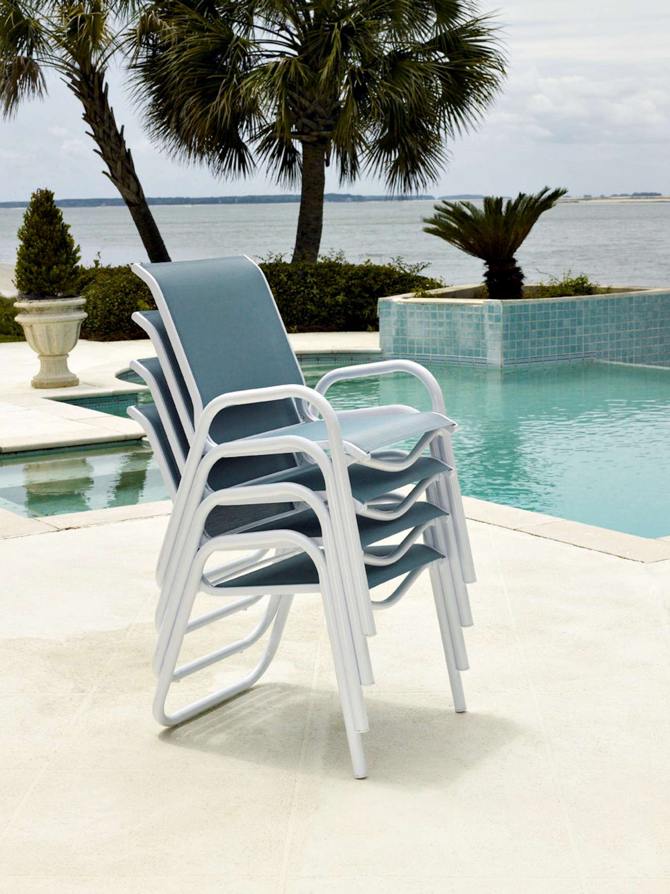 Stack of white plastic chairs beside an infinity pool overlooking a calm sea, with overcast skies and a fire pit nestled among tropical trees in the background.