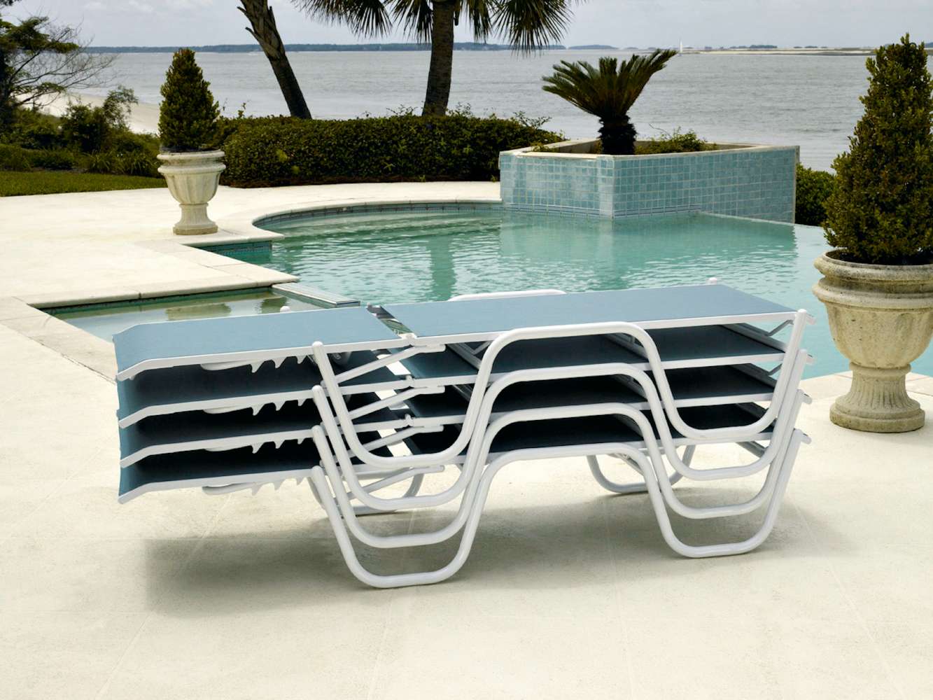 A unique bone-shaped, white and blue lounge chair beside an outdoor infinity pool overlooking a tranquil lake, with lush greenery and a fire pit in the background.
