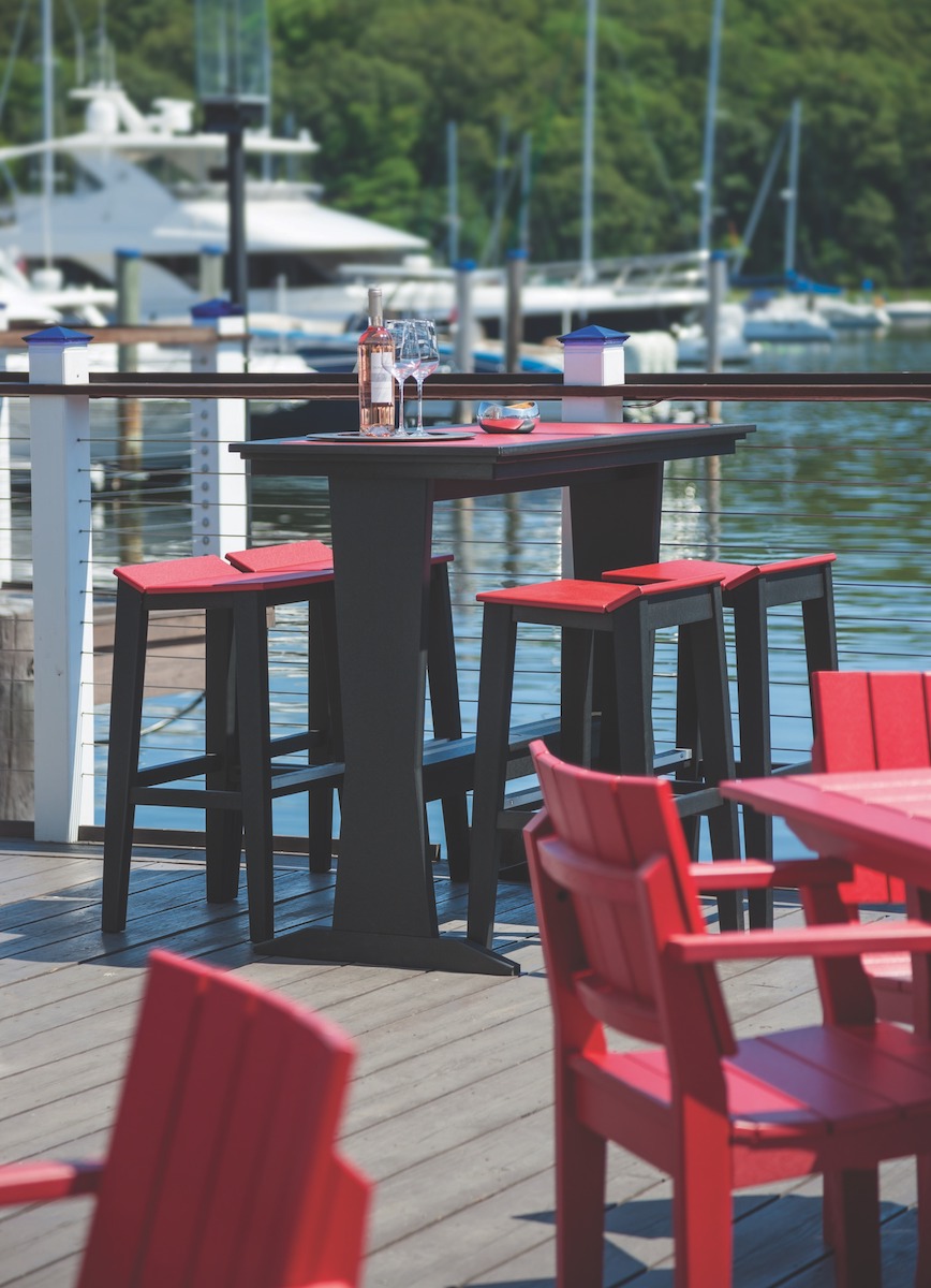 An outdoor bar setup beside a marina with a high-top black table and stools. There are a water bottle and sunglasses on the table, surrounded by lush greenery and boats in the distance, with