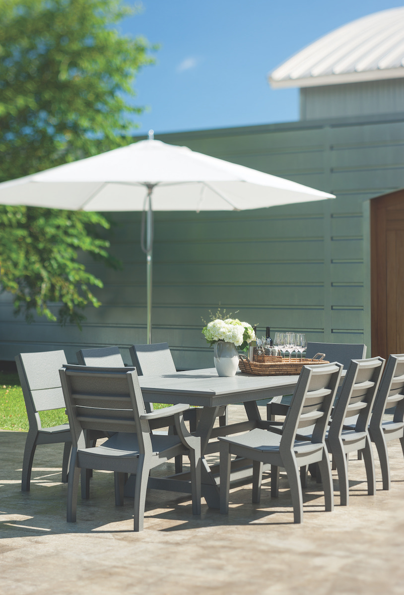 Outdoor dining setup with a grey table and chairs under a white umbrella, framed by green grass and a wooden building under a bright blue sky, featuring an elegant fireplace insert.