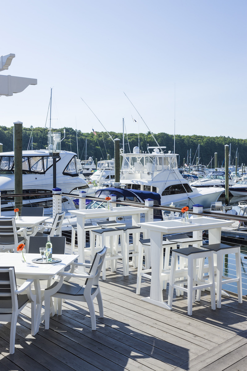 Outdoor dining area with white tables and chairs on a wooden deck overlooking a marina filled with boats under a clear blue sky, enhanced by a cozy fire pit.