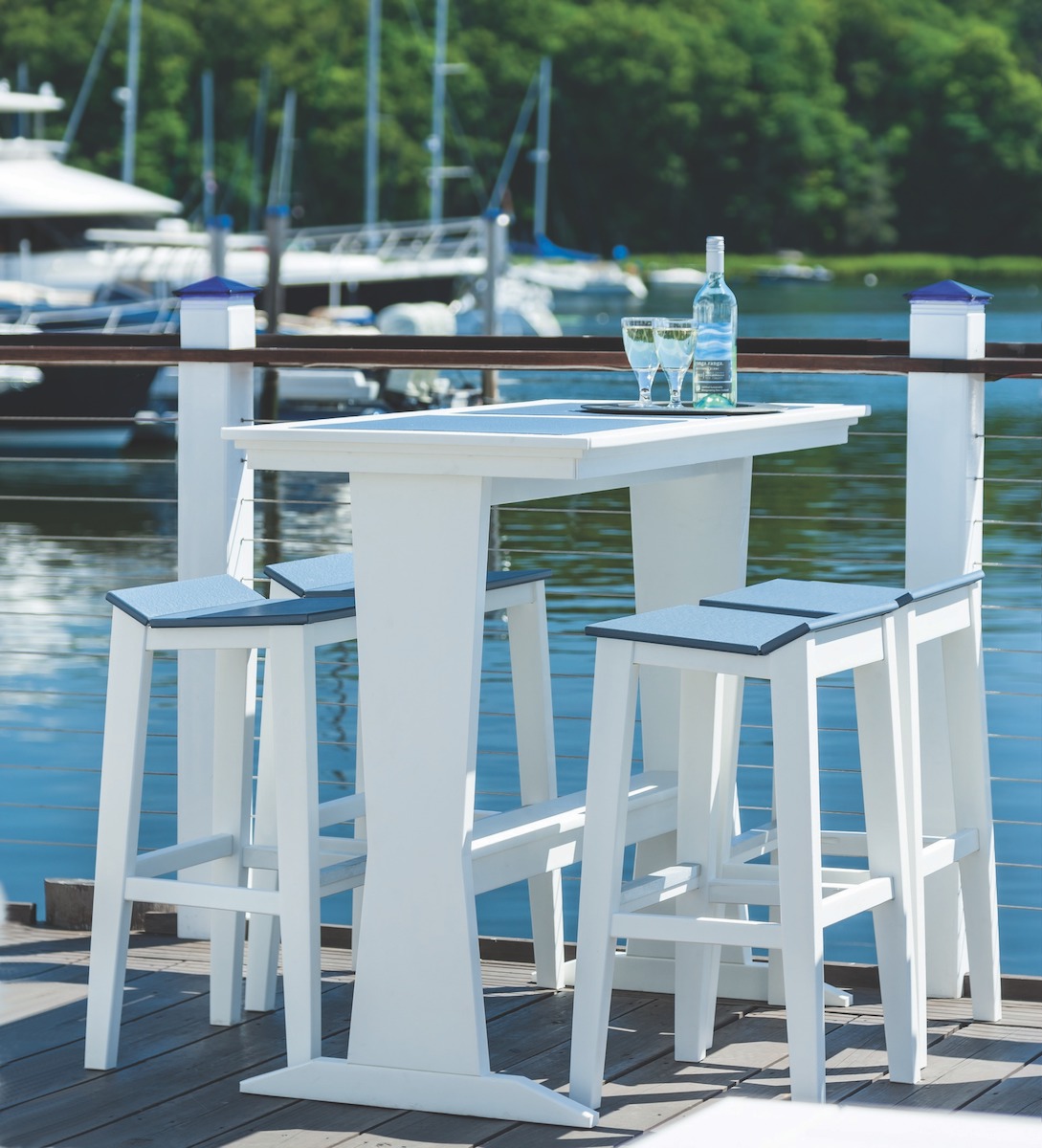 White high-top table with two stools set on a dock, overlooking a marina with boats. A bottle and two glasses sit on the table, next to an elegant fireplace insert, suggesting a relaxing scene