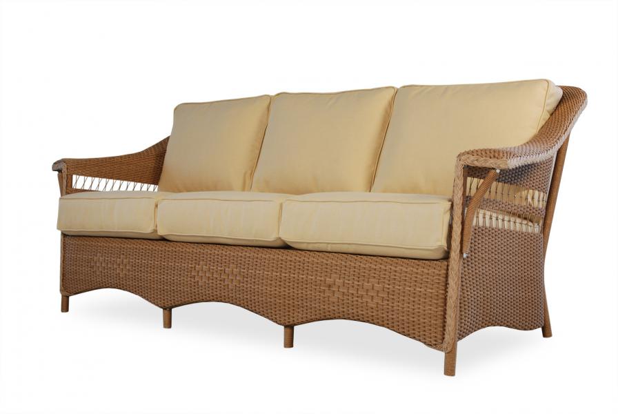 A wicker three-seater sofa with light beige cushions, isolated on a white background. The sofa features a gently curved back and armrests with a woven texture, perfect for placement near a fireplace