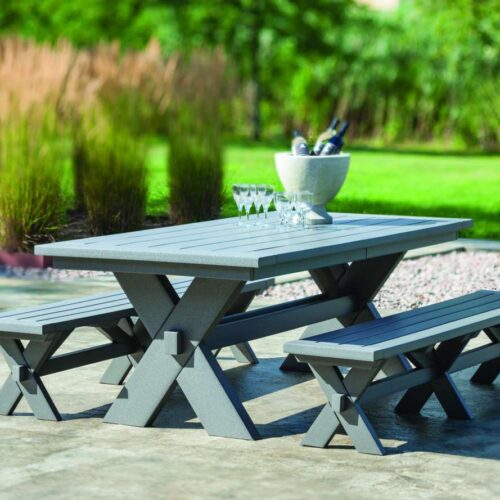 A modern gray picnic table with two benches set on a paved surface near a cozy fire pit, surrounded by a lush green garden with tall grasses. A wine glass and a wine bucket are placed on