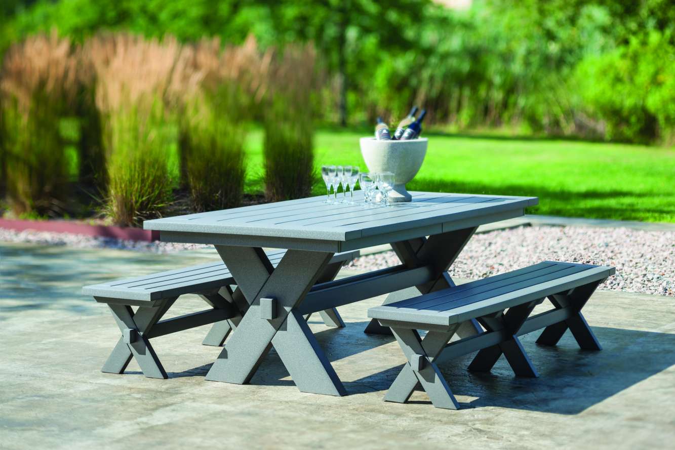 A modern gray picnic table with two benches set on a paved surface near a cozy fire pit, surrounded by a lush green garden with tall grasses. A wine glass and a wine bucket are placed on
