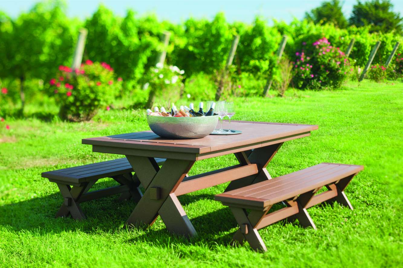 A wooden picnic table with attached benches is set on a lush green lawn, with a bucket of drinks and a fire pit in the background. Vibrant flowers bloom on the side.
