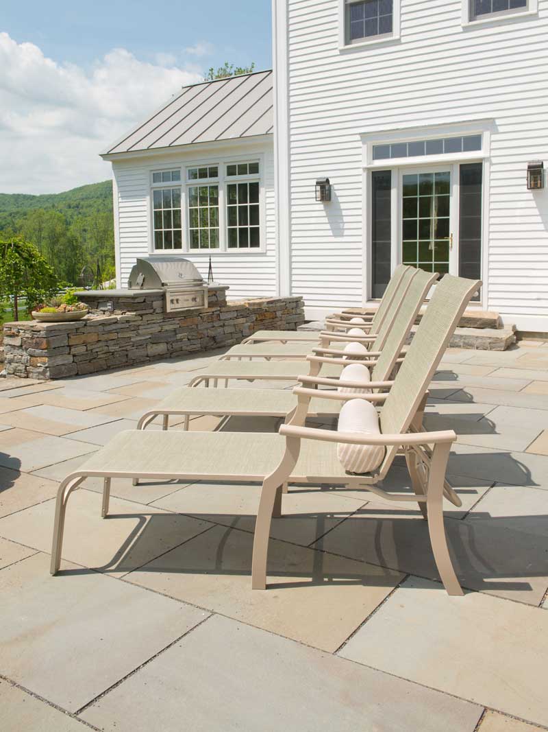 Three beige lounge chairs on a patio with beige tiles, located beside a white house with a mountain landscape in the background and a fireplace insert on a sunny day.