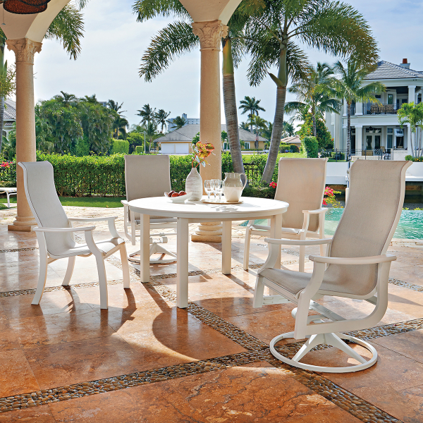 Outdoor patio dining setup with a white table and chairs overlooking a pool and waterfront estate, situated under palm-shaded colonnade with an insert fireplace.