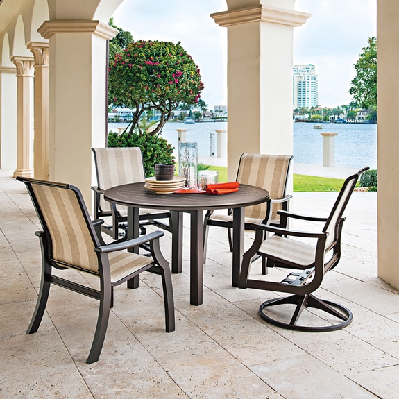 An outdoor dining setting on a veranda featuring a rectangular table and four chairs overlooking a scenic lake, with columns, a flowering bush, and an insert fireplace nearby.