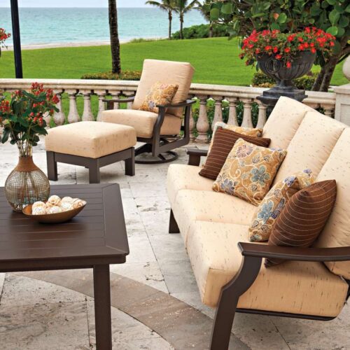 An elegant outdoor seating area with a plush beige sofa and chairs, a brown coffee table, decorative cushions, and a scenic view of a lush green lawn and ocean in the background, complete with a cozy