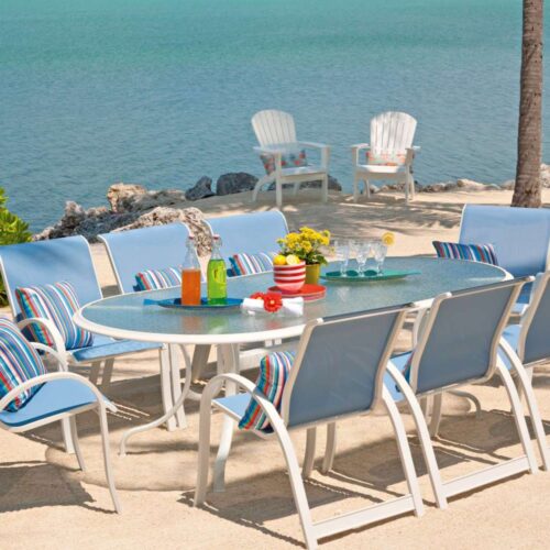 A seaside outdoor dining setup with a glass table surrounded by blue and white chairs, and drinks on the table, featuring a fireplace insert overlooking the ocean and framed by palm trees.
