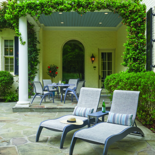 Two lounge chairs and a small table with a book and drink are positioned on a stone patio under a covered archway, next to an outdoor stove, surrounded by lush ivy and a traditional white brick