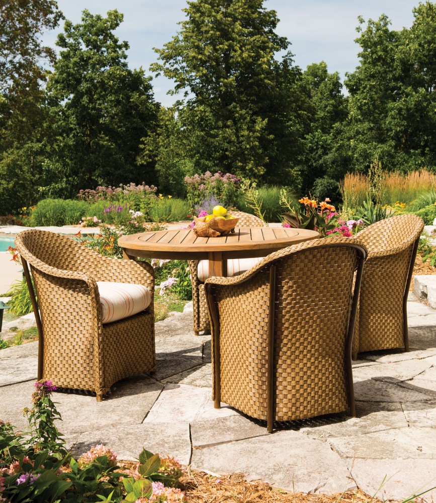 An outdoor dining area featuring a round table and four wicker chairs on a stone patio, surrounded by lush gardens and a distant pool under a clear sky, complete with a cozy fire pit.