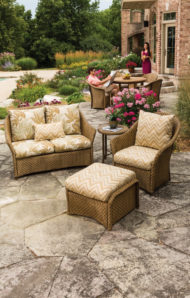 Two people relaxing in a lush garden patio setting with a woman standing by a table serving drinks and a man lounging on a sofa, surrounded by colorful flowers, stylish outdoor furniture, and an elegant fireplace