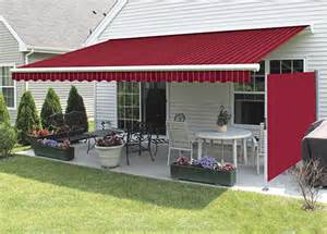 A spacious backyard featuring a red-striped awning extended over a patio with a dining set and a pizza oven, surrounded by neatly landscaped grass and shrubs near a white house.