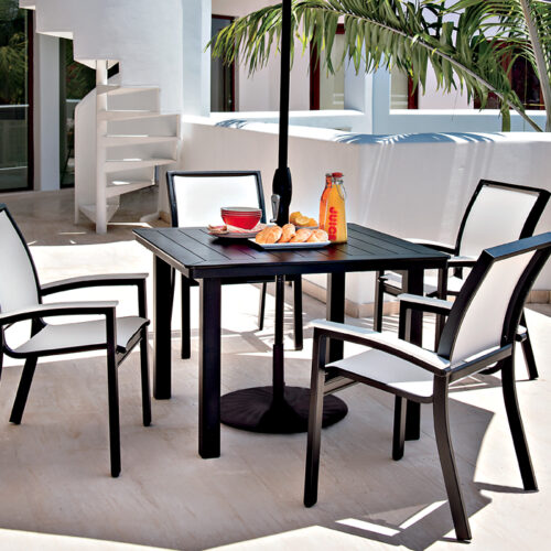 A modern outdoor dining setup on a white patio featuring a black table with four chairs, and breakfast items including a bottle of juice, a bowl of fruits, and an insert grill.