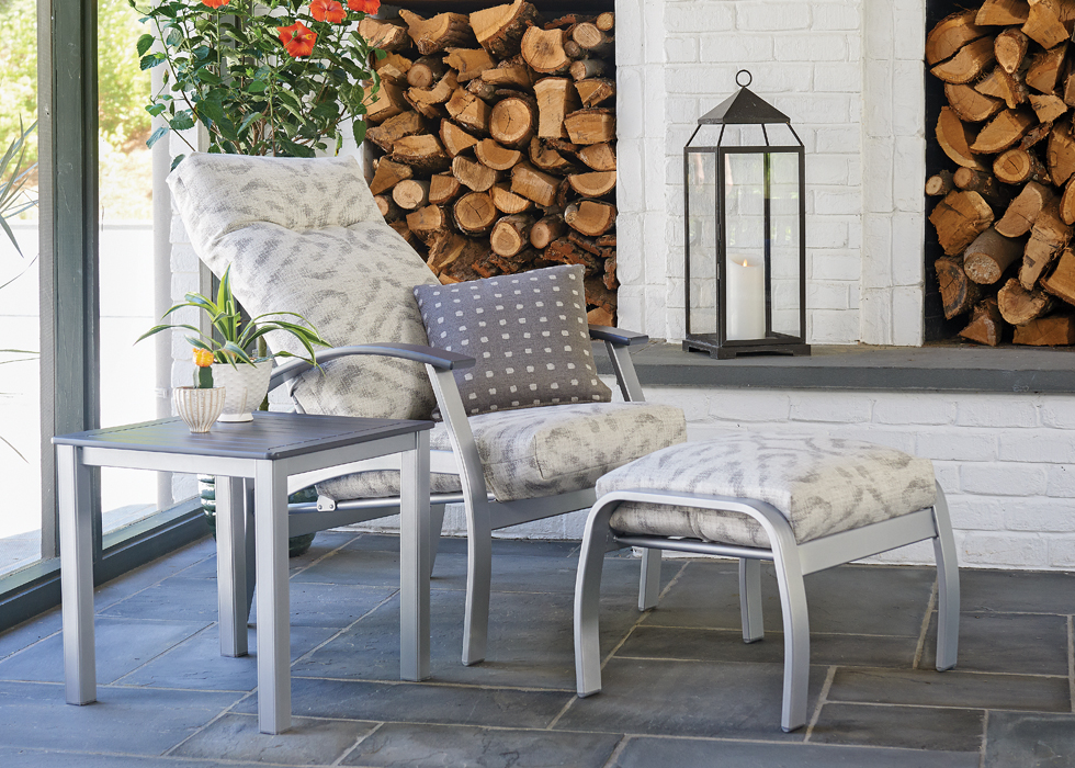 Cozy patio setup with a cushioned lounge chair and side table, a lantern on the table, and a fire pit in the background near flowering plants.