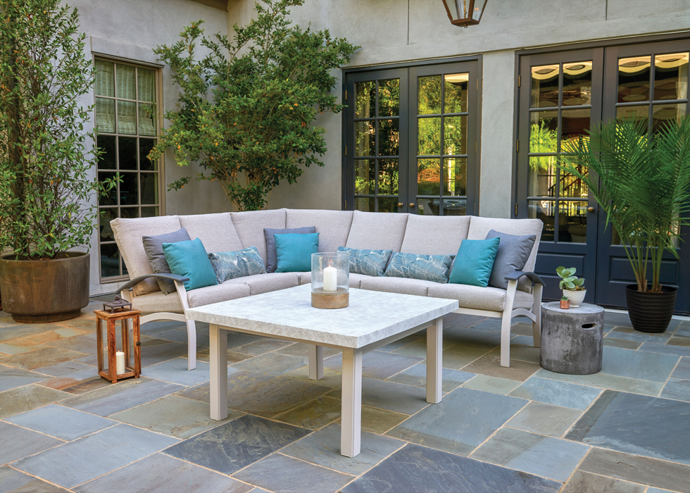 A cozy outdoor patio area featuring a white corner sofa with teal and light blue cushions, a white marble table, and various potted plants, set on a stone-tiled floor with a backdrop of green