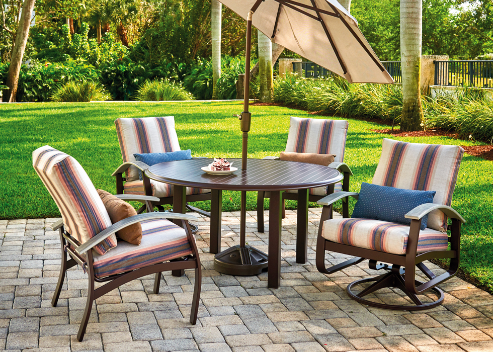 Outdoor patio with four striped cushion chairs, a round table with a snack, and a large umbrella, set on a brick floor with lush greenery in the background and a cozy fire pit nearby.