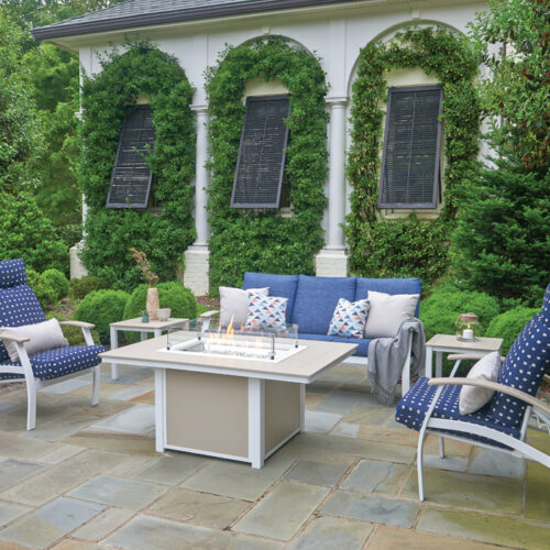 An elegant outdoor seating area with a white sofa, two chairs with blue cushions, and a coffee table, against a backdrop of white columns, a cozy fireplace, and lush greenery.