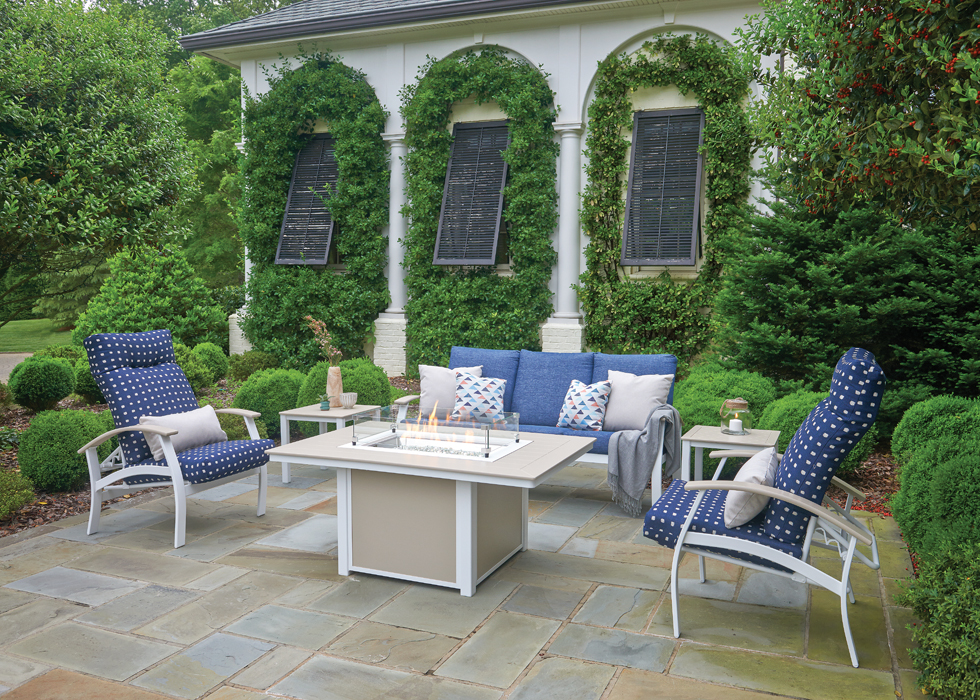 An elegant outdoor seating area with a white sofa, two chairs with blue cushions, and a coffee table, against a backdrop of white columns, a cozy fireplace, and lush greenery.