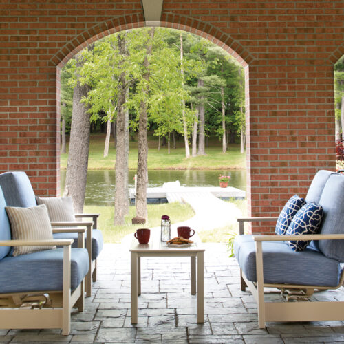 Four chairs on a porch with cushions, overlooking a tranquil lake surrounded by trees, seen through a brick archway. A table holds books and a cup next to a cozy fire pit, suggesting a relaxing