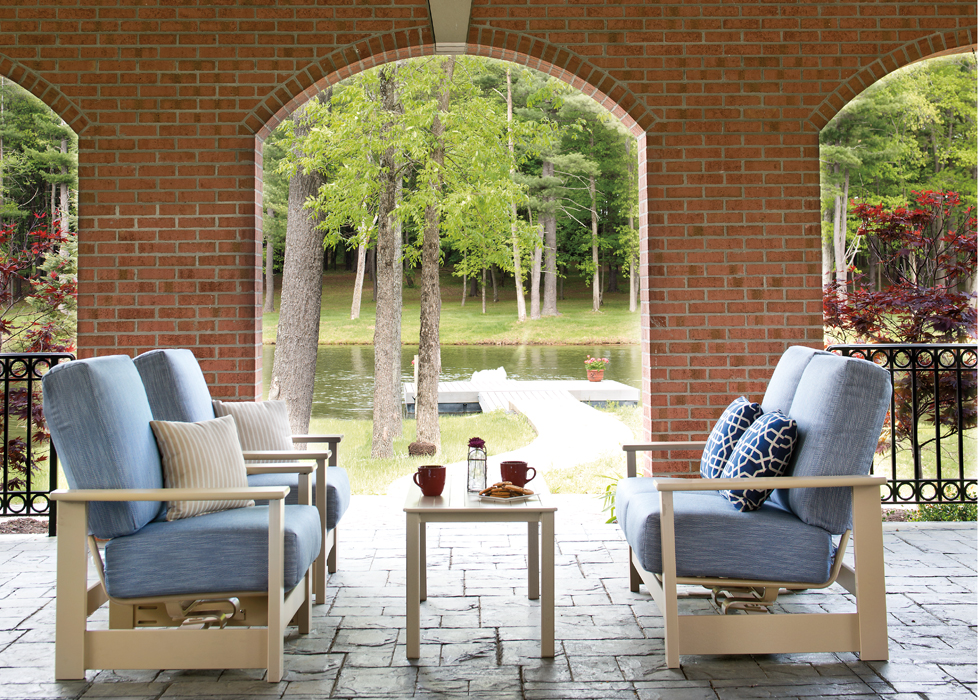 Four chairs on a porch with cushions, overlooking a tranquil lake surrounded by trees, seen through a brick archway. A table holds books and a cup next to a cozy fire pit, suggesting a relaxing