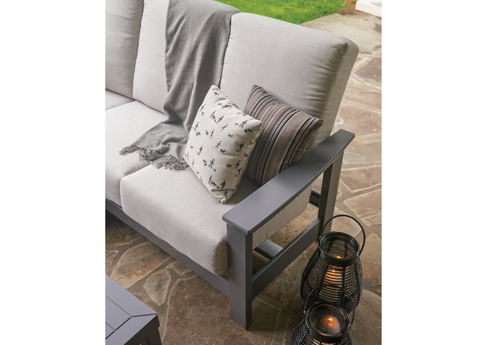A modern outdoor sofa with gray cushions, featuring an integrated black side table and decorative pillows, placed on a stone patio next to a fire pit and lantern-style candle holders.