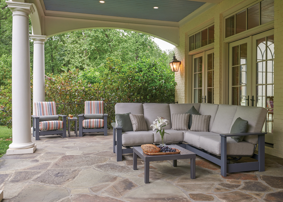 An elegant porch with stone flooring, featuring a comfortable seating area with a gray sofa and chairs with striped cushions, a small table with a fire pit, surrounded by lush greenery.
