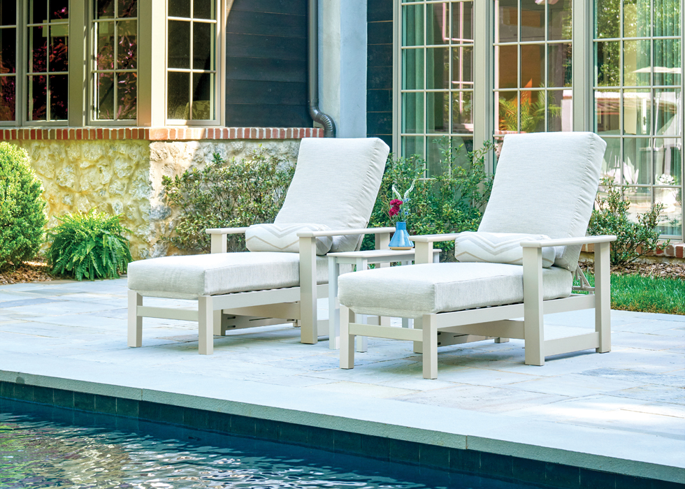 Two white lounge chairs with cushions next to a small ottoman, arranged neatly by a poolside, with a fire pit and greenery in the background.