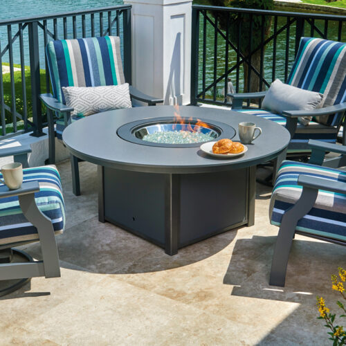 Outdoor patio with a fireplace table surrounded by four striped cushioned chairs, overlooking a body of water, with a breakfast tray on the table.