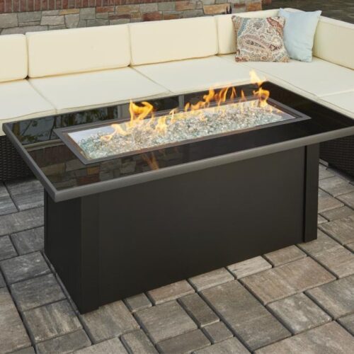 A modern outdoor gas fire pit with bright flames, surrounded by a black metal frame on a stone-paved patio, complemented by a cream cushioned corner sofa.