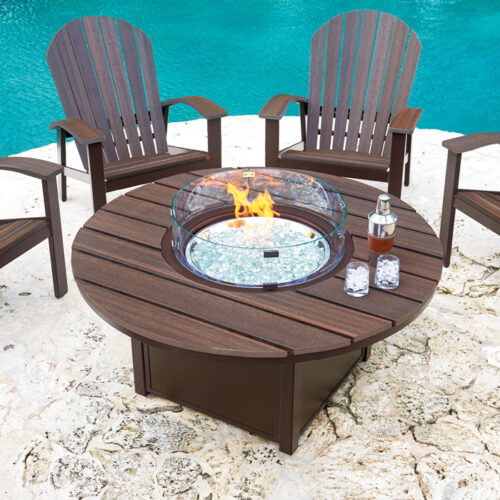 An outdoor patio setup with a circular, wooden fire pit insert table surrounded by four dark brown adirondack chairs, on a white pebbly surface near a clear blue swimming pool.