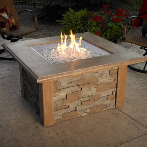 A square fire pit with a lit flame, surrounded by glass beads, is set on a patio. Nearby are two chairs with patterned cushions, and a flowerbed with red blooms enhances the cozy outdoor