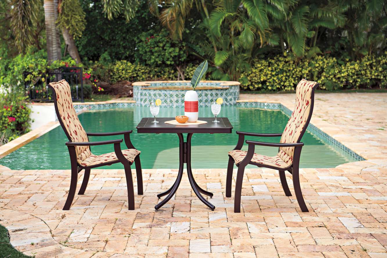 Two ornate chairs and a small table set by a pool, surrounded by lush greenery. A red candle and a book are on the table, close to an outdoor fire pit.