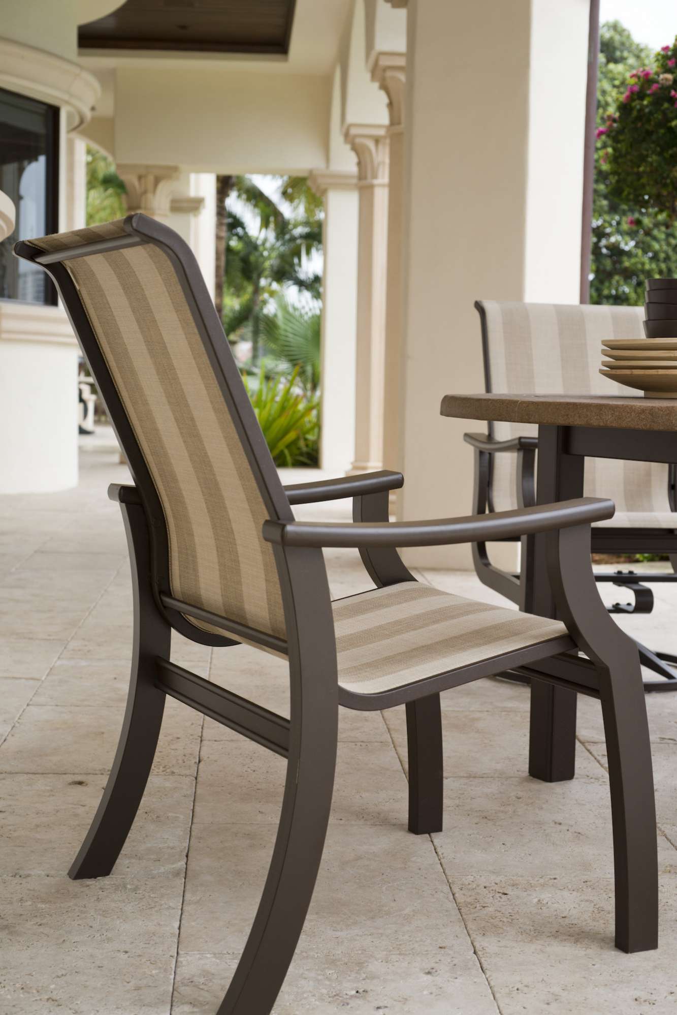 A comfortable outdoor dining chair with a metal frame and striped upholstery, set on a patio with a fireplace, beige columns, and tiled flooring, hinting at a luxurious, warm setting.