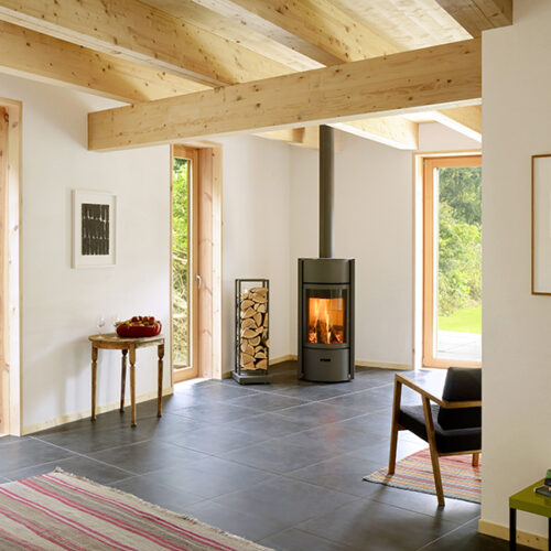 Bright, modern living room with exposed wooden beams, a fire pit, and large doors opening to a sunny garden. Decor includes framed artwork, a striped rug, and minimalist furniture.